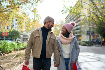 Beautiful young couple interracial in love enjoying winter holiday season in the city streets, taking a walk, hugging and holding gift bags while celebrating Christmas outdoors