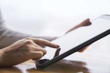 Poster - Woman's fingertip gently tapping the screen of a modern digital tablet lying on a desk, surrounded by the unfocused brightness of a contemporary office space