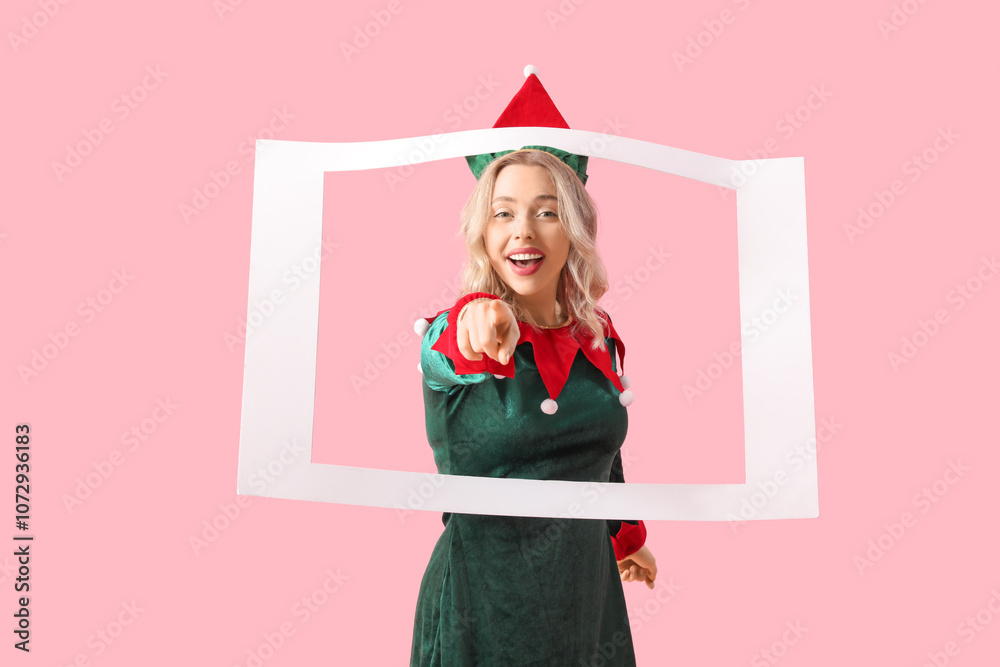 Beautiful young woman dressed as elf with frame pointing at viewer on pink background