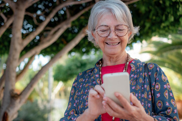 Wall Mural - Smiling senior woman in public park while using mobile phone, caucasian elderly modern lady enjoying tech and social