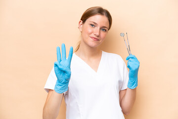 Wall Mural - Dentist caucasian woman holding tools isolated on beige background happy and counting three with fingers