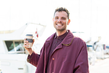 Wall Mural - Young caucasian man holding a take away coffee at outdoors smiling a lot