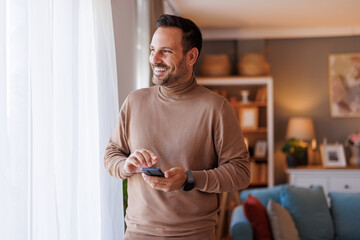 Wall Mural - Happy mid adult man with mobile phone looking through window thoughtfully while standing in living room