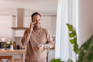 Wall Mural - Cheerful mid adult man looking through window and talking over phone call while standing in home office