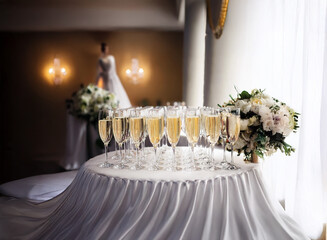 A table with champagne flutes and white flowers sits in a room with a wedding dress in the background.