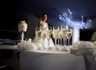 Poster - A table with a white tablecloth is set with champagne flutes, the bride is standing in the background, the room is dimly lit with white string lights.