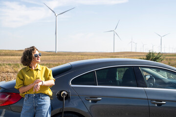 Wall Mural - Woman with curve hairs wearing yellow shirt and sunglasses stands next to charging electric car. Wind turbines in the background.