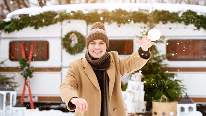 Wall Mural - A young man in warm winter clothing joyfully throws a snowball in a snowy landscape. He stands in front of a decorated camper, surrounded by holiday decorations and trees.