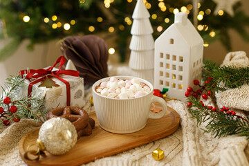 Wall Mural - Warm cup of cocoa with marshmallows and chocolate cookies on wooden tray, christmas ornaments and gift on cozy knitted background against stylish christmas tree with lights. Winter mood