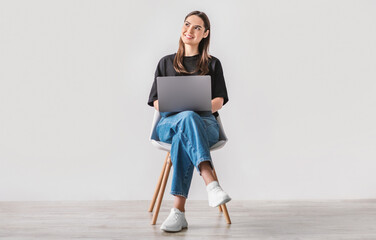 Poster - Beautiful young woman sitting on chair, working on laptop against white studio wall, full length. Dreamy millennial lady using portable pc for work, entertainment or online studies