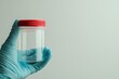 © zakiroff - A hand in a blue glove holds a clear specimen jar with a red lid against a light gray background with copy space