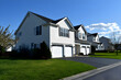 © Emons - Neighborhood Real Estate Image - A row of suburban houses with garages and well-maintained lawns under a clear blue sky.