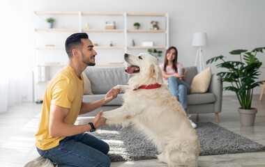 happy young arab guy playing with dog, having fun on floor at home, his girlfriend drinking coffee o