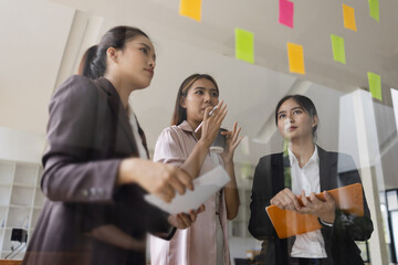 Wall Mural - Asian businesswoman teamwork planning with sticky notes and new ideas on a glass wall, Business people brainstorming strategy and new ideas concept, startup worker meeting,