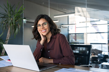 Portrait 30s latin hispanic business woman manager working on laptop computer in modern office. Smiling Indian young businesswoman professional entrepreneur using pc app at workplace looking at camera