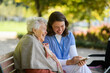 © Halfpoint - Portrait of nurse and elderly woman on walk in the park during warm autumn day. Young caregiver looking through photos of senior patient's family on a phone.