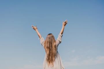 Wall Mural - A woman with long hair is standing on the beach, looking up at the sky. She is wearing a dress and she is happy.