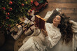 © polinaloves - Young woman reading book, lying near Christmas tree in a cozy decorated room.