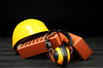 Wall Mural - Red bricks, yellow hard hat and earmuffs on dark wooden table. Building material and safety equipment