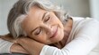 © Liana - Peaceful senior woman with gray hair resting her head on her hands, eyes closed, wearing a white sweater. The serene pose highlights relaxation, comfort, and contentment in a cozy, natural setting.