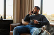 © Dmytro Hai - African american man talking on smartphone and holding coffee cup on armchair at home