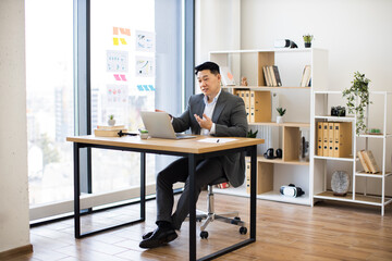 Wall Mural - Asian male businessman in his 30s engaged in online video call while working on laptop in modern office. Professional environment with charts on wall and organized workspace.