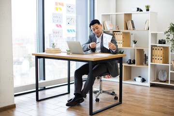 Wall Mural - Asian businessman wearing headset in office showing graphs during online video call. Professional setting with laptop, modern interior, and large window. Business attire suggests formal meeting.