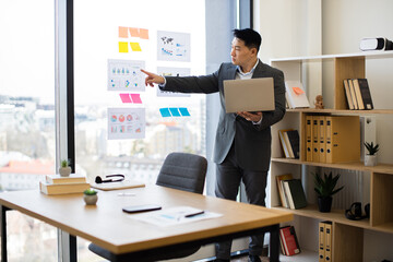 Wall Mural - Asian businessman in professional suit analyzing charts and graphs during video call. Standing by panoramic window with laptop, pointing at data visuals. Modern office setting with urban view.