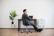 © sofiko14 - Asian man in business attire sitting in office chair, relaxing in modern workspace. Minimalist office features wooden desk, laptop, indoor plant on white wall backdrop.