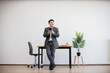 © sofiko14 - Asian businessman in suit uses smartphone while leaning against office desk in minimalist setting. Young professional stands confidently in bright, modern workspace.