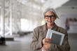 © Ivan Traimak - Stylish elderly woman holding a notebook smiles confidently while standing in a modern architectural space during daylight