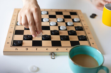 A hand moves a white checker piece on a wooden board during a game. Two coffee mugs, one teal and one yellow, sit nearby. The scene highlights focus, strategy, and a relaxed, casual indoor setting.