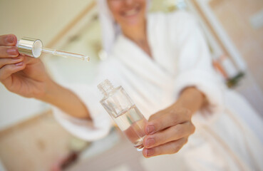 Wall Mural - A woman in a bathrobe and towel on her head uses a dropper to apply skincare serum from a glass bottle. The bright bathroom setting suggests a fresh, self-care beauty routine and wellness lifestyle.