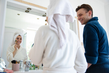 Wall Mural - A woman in a bathrobe with a towel on her head is smiling while using a face roller in front of a mirror. A man in a bathrobe stands beside her, smiling, enjoying a relaxed morning together.