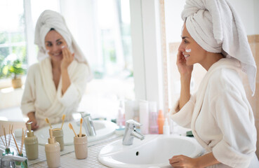 Wall Mural - Smiling woman in white robe applies skincare cream while looking at herself in bathroom mirror. Her hair is wrapped in towel, surrounded by beauty products and plants, evoking fresh, relaxing vibe