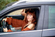 © Connect Images - Red-haired woman smiling while driving a car with a passenger in the backseat.