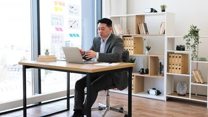 Wall Mural - Asian businessman in his 30s wearing headset during online video call in modern office. He sits at desk with laptop, gesturing while speaking.