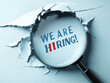© wutipong - a magnifying glass on a light blue background. The magnifying glass is revealing the words 'We Are Hiring!' through a hole in the paper. The words are written in blue and red, with in 'Hiring'