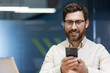 © Tetiana - Close-up photo of a young businessman with glasses and a beard sitting in the office at his workplace and using a mobile phone