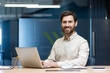 © Tetiana - Portrait of a smiling young male office worker sitting at a desk, working on a laptop, looking confidently into the camera