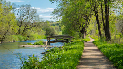 Wall Mural - A riverside trail with fresh greenery on both sides, and a small wooden bridge crossing over the river, offering a peaceful setting for running or nature walks.