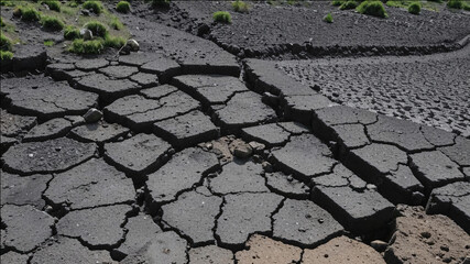 Close-up of cracked, dry mud in a barren landscape, showcasing dark, fractured earth with scattered patches of green moss and distant blurred rocks under dramatic lighting.
