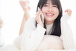 © kapinon - うつ伏せでスマホを通話するニットを着た若いかわいい笑顔の女子のクローズアップ　明るい白いかわいいコミュニケーションなどのイメージ