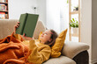 © Westend61 - Smiling girl reading book and lying with knitted blanket on sofa at home