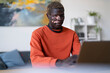 © Studio Marmellata - A young man with vitiligo dressed in an orange shirt types on a laptop while seated at a desk in a bright office with modern furniture and artwork, surrounded by a relaxed and professional atmosphere.