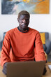 © Studio Marmellata - man with vitiligo wearing an orange shirt sits at a desk, smiling warmly in a modern home office setting with colorful artwork and decor, embodying a relaxed and positive ambiance