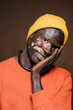 © Studio Marmellata - Black man with vitiligo wearing a yellow beanie and orange shirt rests his face on his hand, smiling softly with a relaxed and content expression against a warm brown background.