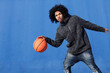 © Raul Mellado - african american teen male in informal outfit playing basketball with ball near blue background looking at camera, concept of sport and young lifestyle, copy space for text