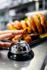 Wall Mural - A hand reaching for a service bell in a restaurant setting, with a blurred background featuring a sandwich and fries. Ideal for hospitality, customer service, or dining-related themes.