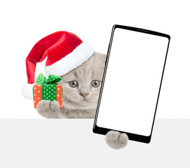 Aufkleber - Cute kitten wearing red christmas hat holds tiny gift box and  smartphone with white blank screen in it paw looks above blank white banner. Empty space for text. Isolated on white background